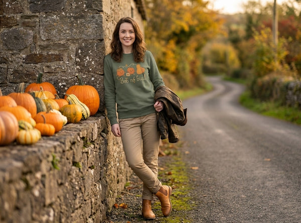 Woman wearing the olive Bob nó Bia adult sweatshirt leaning on a stone wall stacked with pumpkins