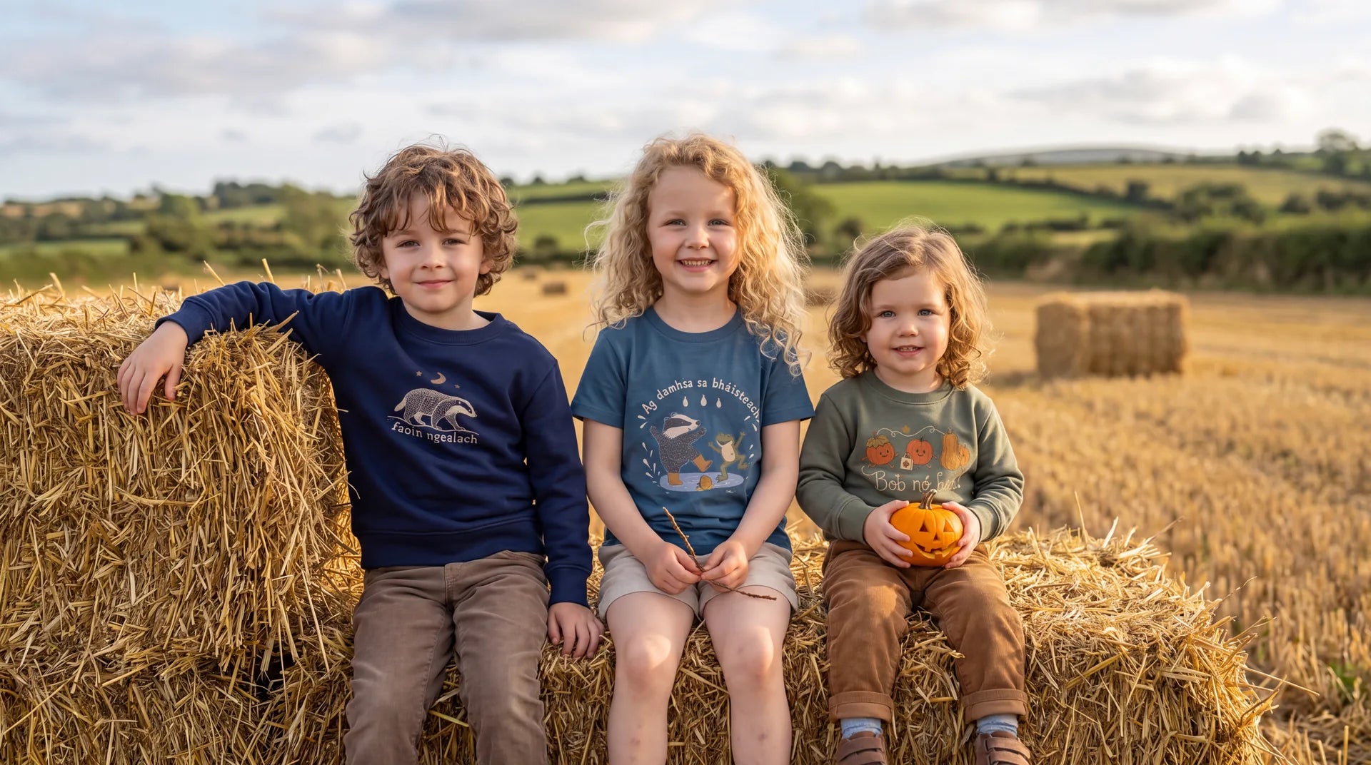Three children on a hay bale in a harvested Irish field wearing Glasóg autumn designs, one holding a small carved pumpkin