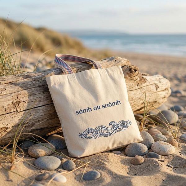 Sámh ar Snámh Irish language tote bag leaning on driftwood on an Irish beach — natural recycled cotton tote with rainbow webbing handles. Glasóg Gaeilge shopping bag.