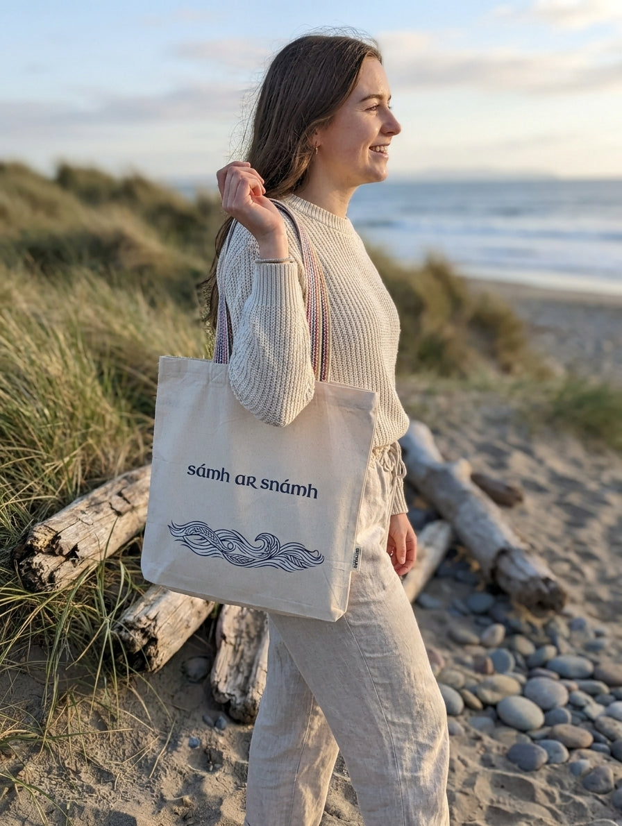 Woman on an Irish beach carrying a Glasóg Sámh ar Snámh recycled cotton tote bag with wave design and rainbow handles. Irish language gift for sea swimmers.