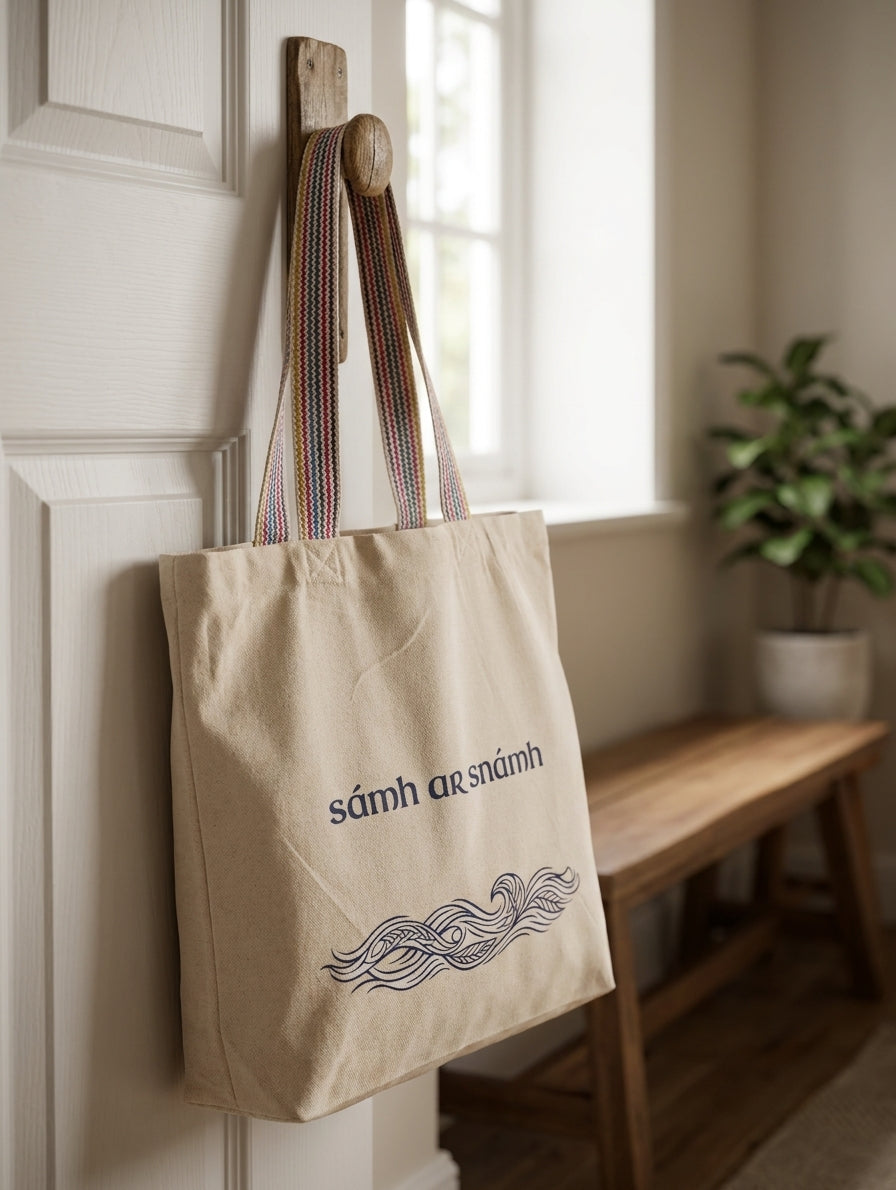 Glasóg Sámh ar Snámh recycled cotton tote bag hanging on a wooden peg indoors — Irish language Gaeilge shopping bag designed in Wexford, Ireland.