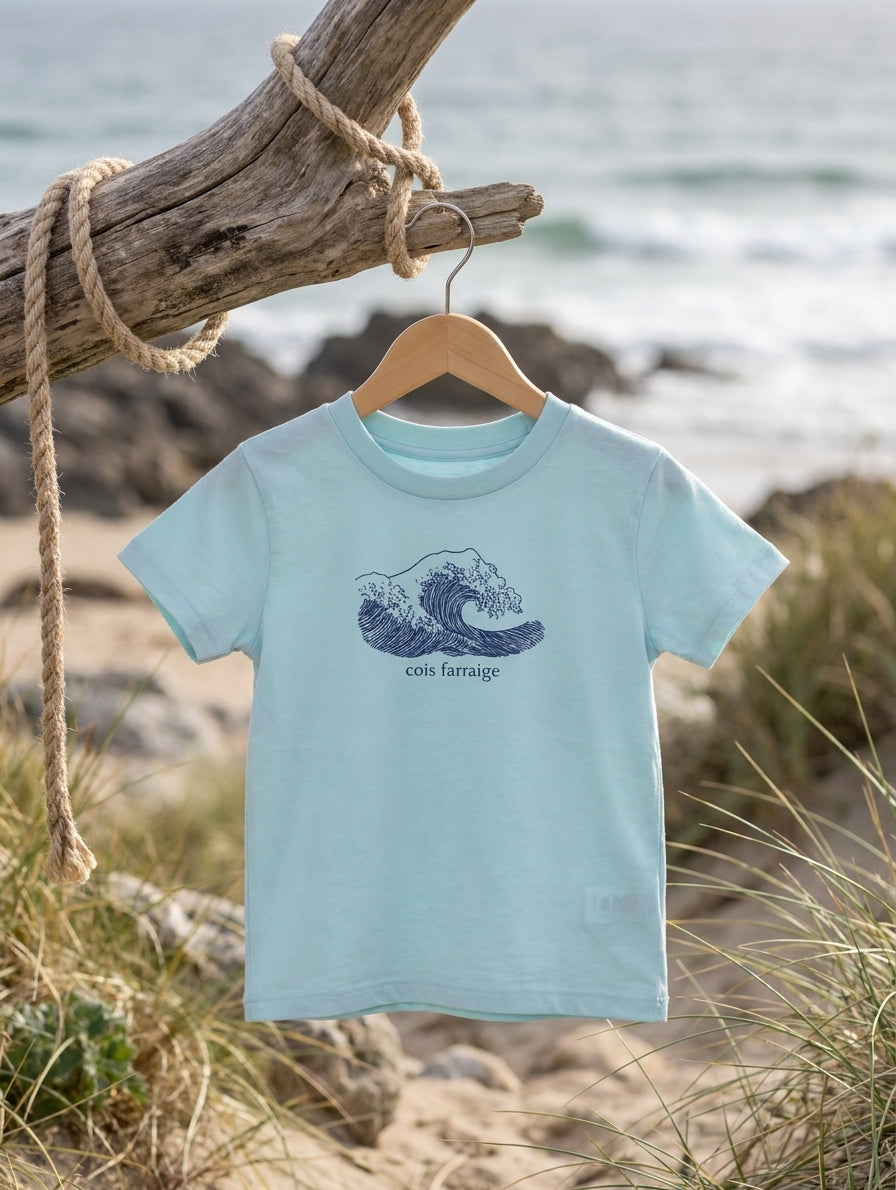 pool blue kids t-shirt with wave illustration hanging on a wooden hanger outdoors by the Irish coast. Glasóg organic cotton Gaeilge clothing.