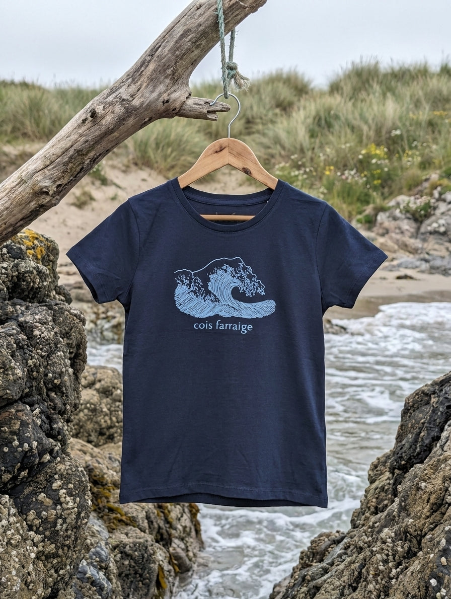 navy kids t-shirt with wave illustration hanging on a wooden hanger outdoors by the Irish coast. Glasóg organic cotton Gaeilge clothing.