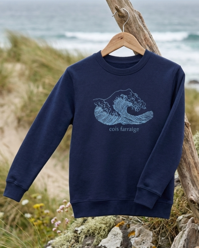 Navy Cois Farraige sweatshirt hanging on a wooden hanger by the Irish coast