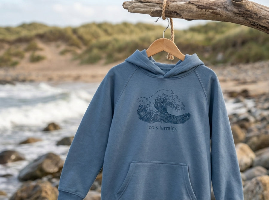 Blue Cois Farraige kids hoodie hanging on a wooden hanger by the Irish coast