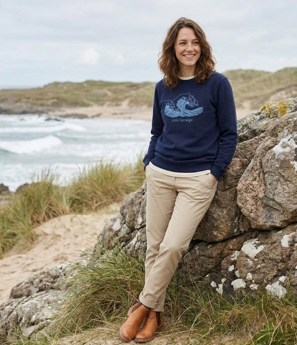 Woman wearing the navy Cois Farraige adult sweatshirt leaning on a rock above the strand