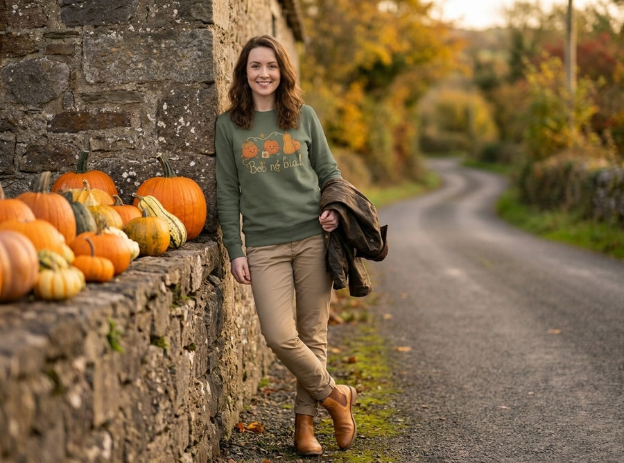 Woman wearing the olive Bob nó Bia adult sweatshirt leaning on a stone wall stacked with pumpkins