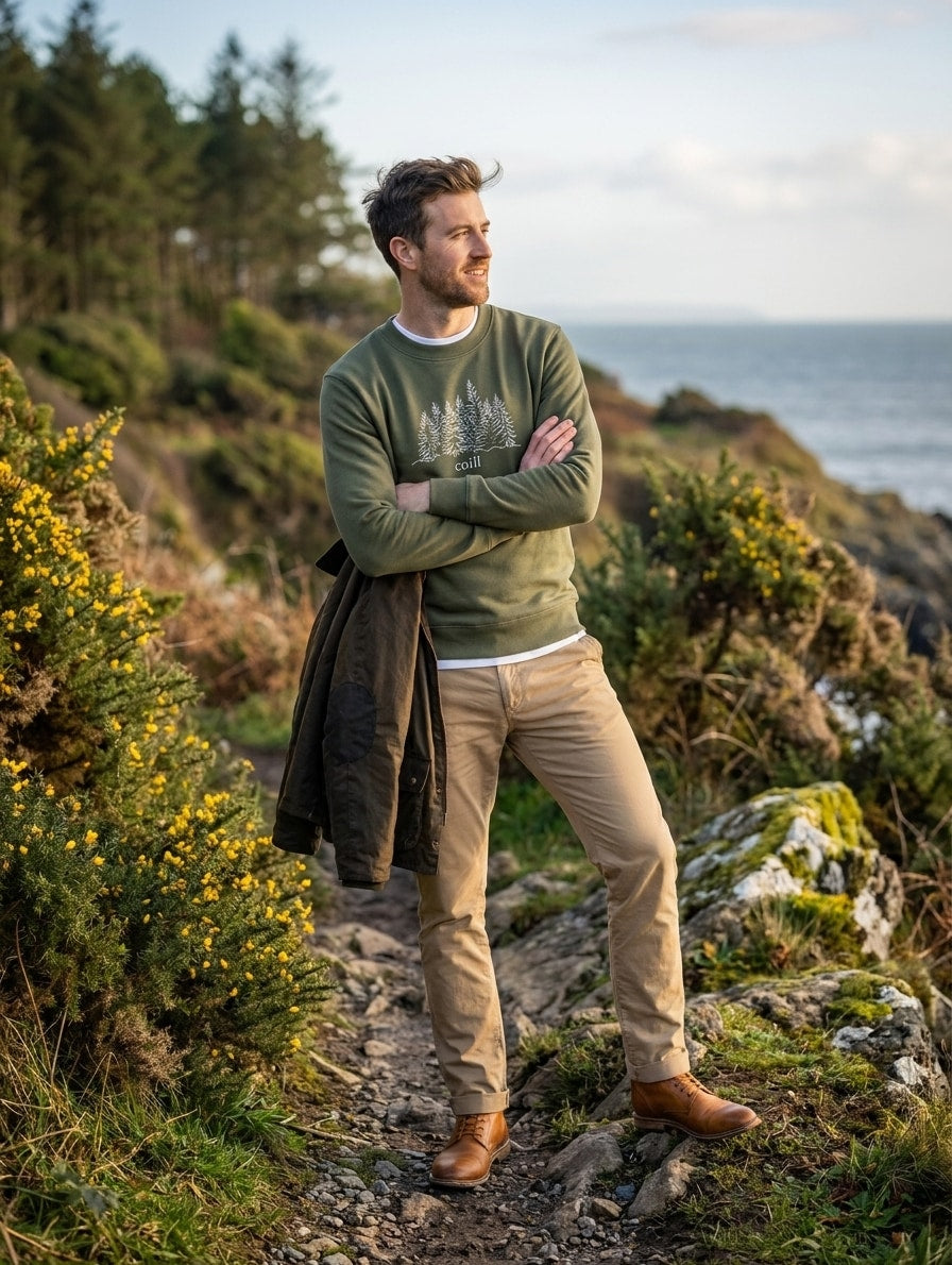 Man wearing the olive Coill adult sweatshirt on a coastal path with gorse and pine woods behind