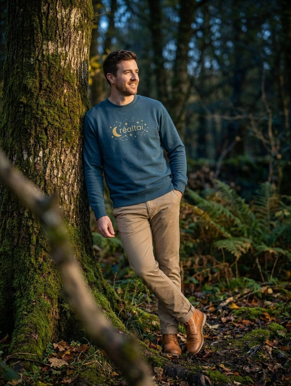 Man wearing the twilight blue Réaltaí adult sweatshirt leaning against a mossy tree in Irish woodland